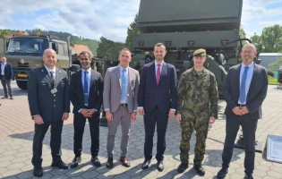 Six men stand outdoors in front of military vehicles and equipment; five wear business suits and one wears a camouflage military uniform, all smiling and posing for the photo on a sunny day.