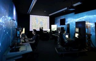 People sit at computer stations in a dark room with large digital maps and blue, network-themed graphics—such as Link16 and NATO data—projected on the walls, suggesting a high-tech operations or control center.