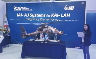 Two men sit at a table signing documents with South Korean and Israeli flags. A woman stands beside them. Behind them is a large banner displaying a helicopter and the text “IAI-AJ Systems for KAI-LAH Signing Ceremony.”.
