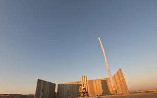 A rocket launches into the clear sky, leaving a white trail behind, as a person sits near a structure made of large metal panels in a barren landscape—an image reminiscent of an IAI annual report.