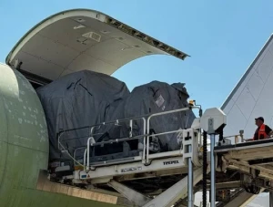 A cargo plane, possibly an IAI conversion, with its nose open is being loaded with large, tarp-covered items using specialized equipment. A worker in a safety vest stands nearby, overseeing the process under a clear sky.