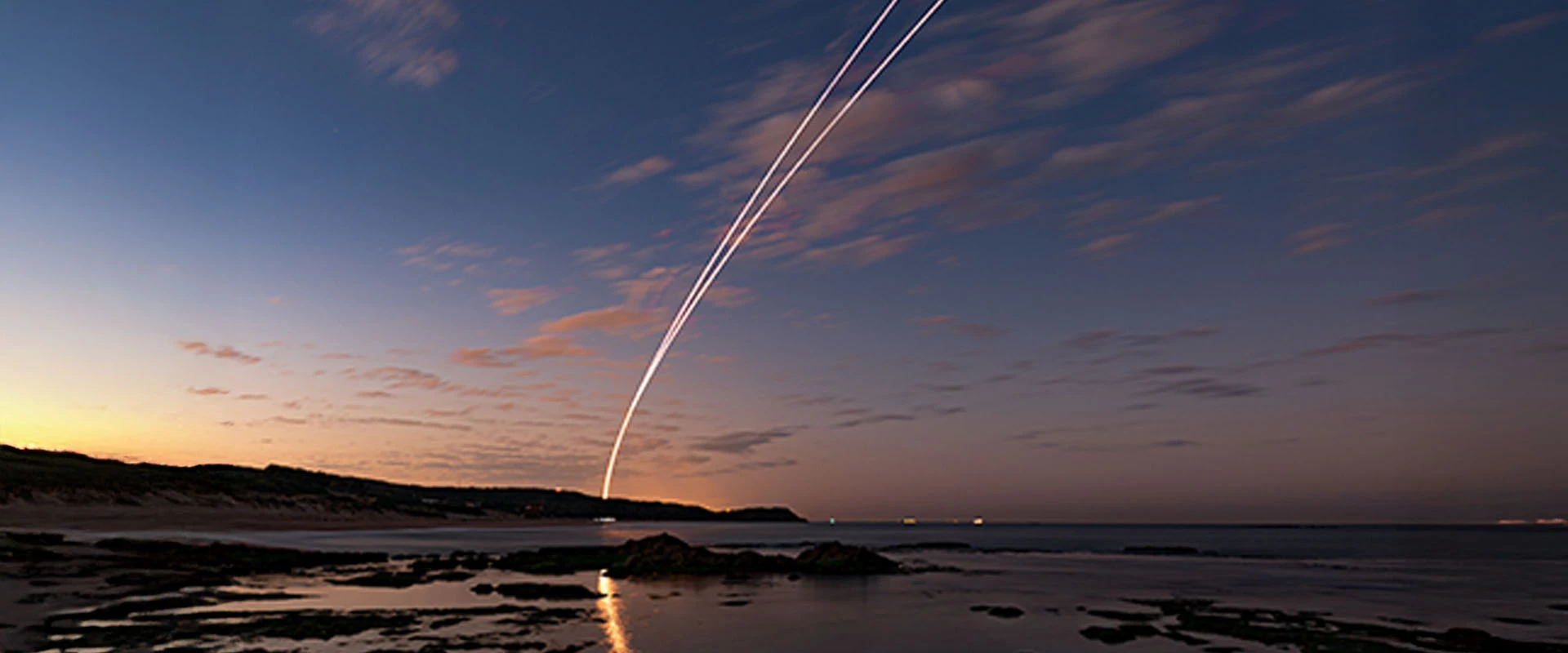 A long exposure photo of a coastal landscape at dusk, with streaks of light arching across the sky—possibly the trail of an anti ballistic missile—reflecting on the calm water below.