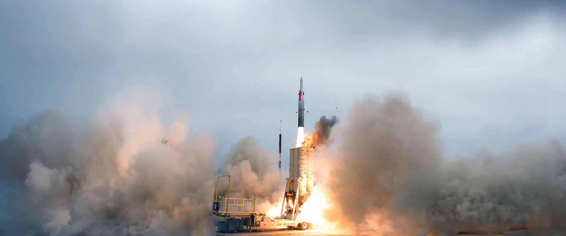 A missile interceptor arrow launches vertically from a ground-based platform, surrounded by thick clouds of smoke and fire, set against a cloudy sky.