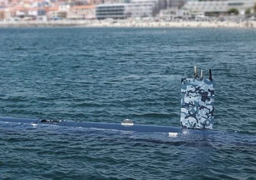 An advanced autonomous submarine with a blue camouflage conning tower is partially surfaced in the water, with a coastline and blurred buildings visible in the background.