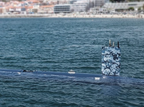 An advanced autonomous submarine with a blue camouflage conning tower is partially surfaced in the water, with a coastline and blurred buildings visible in the background.