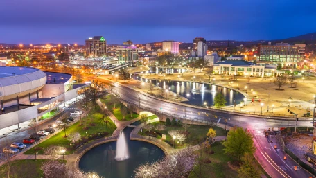 A cityscape at dusk with brightly lit buildings, a domed arena, and winding roads. A fountain flows into a small pond, surrounded by trees and walkways, reflecting the citys lights.