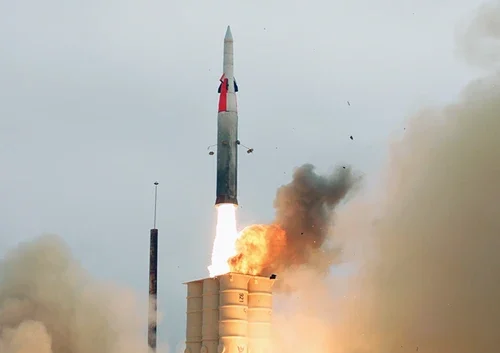 An Arrow missile interceptor is being launched vertically into the sky, with flames and smoke billowing from its base against a cloudy background. The missile emerges from a rectangular launch platform.