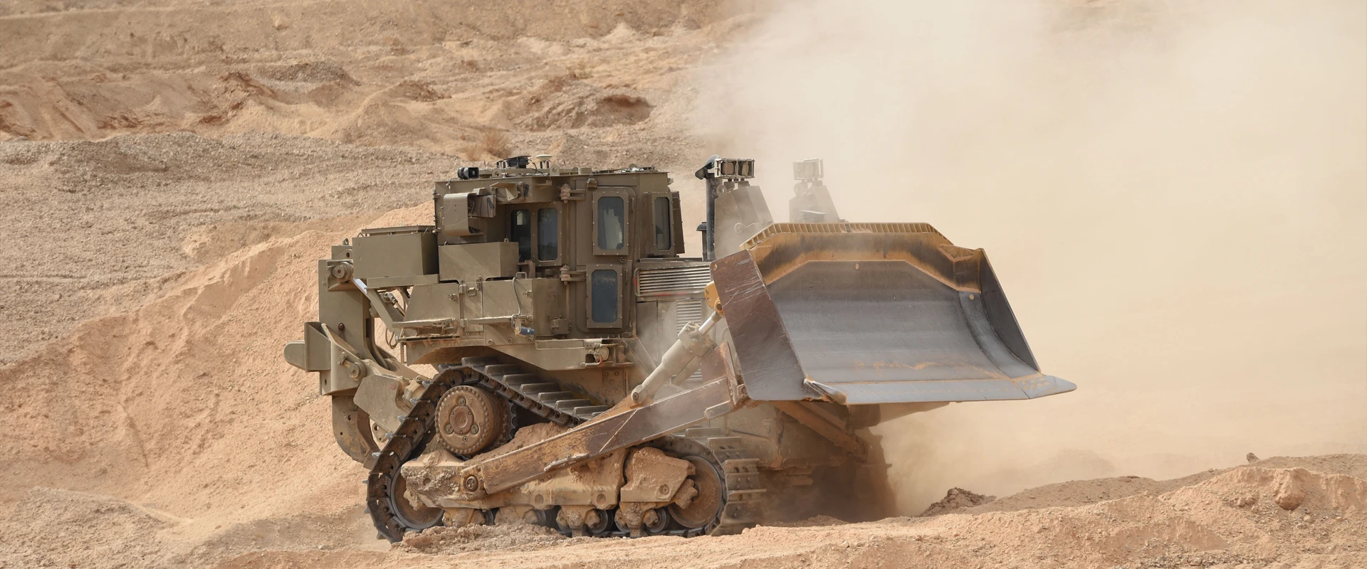 A large, armored bulldozer equipped with an advanced engineering system moves sand and dirt in a desert environment, kicking up dust as it operates on rugged terrain.