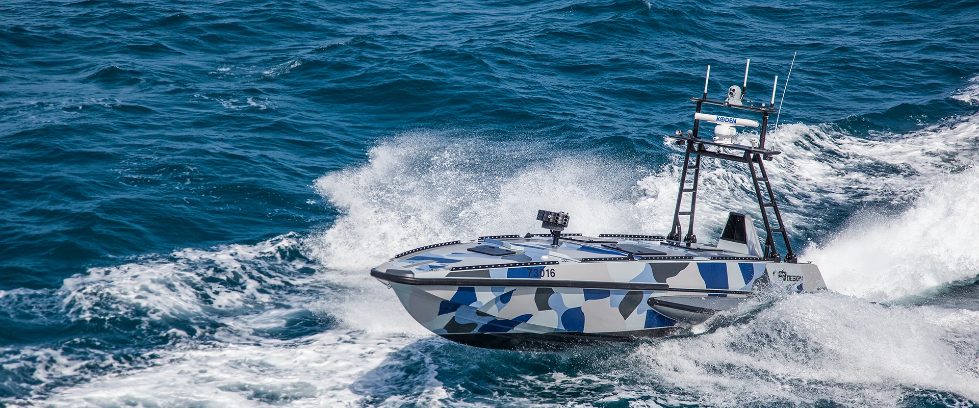 A small, unmanned USV system with a blue and gray camouflage pattern speeds through choppy ocean waters, creating white spray behind it under a clear sky.