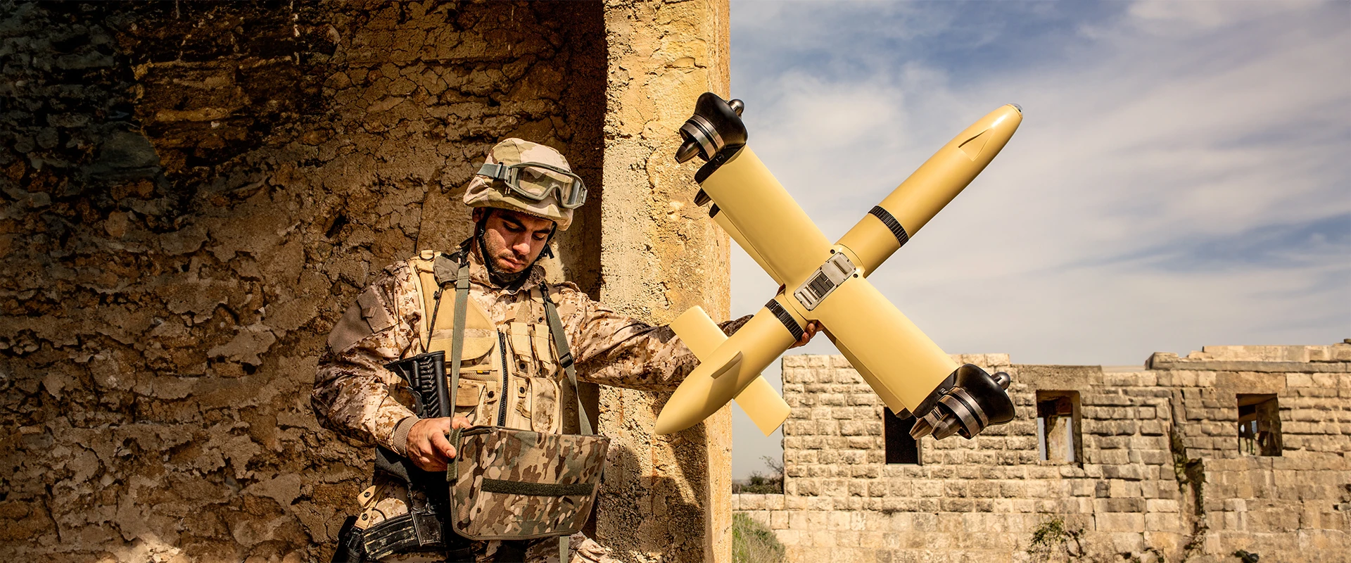 A soldier in desert camouflage operates military equipment beside a weathered building, one hand ready at a missile point, while a yellow drone with four propellers hovers nearby under a blue sky.