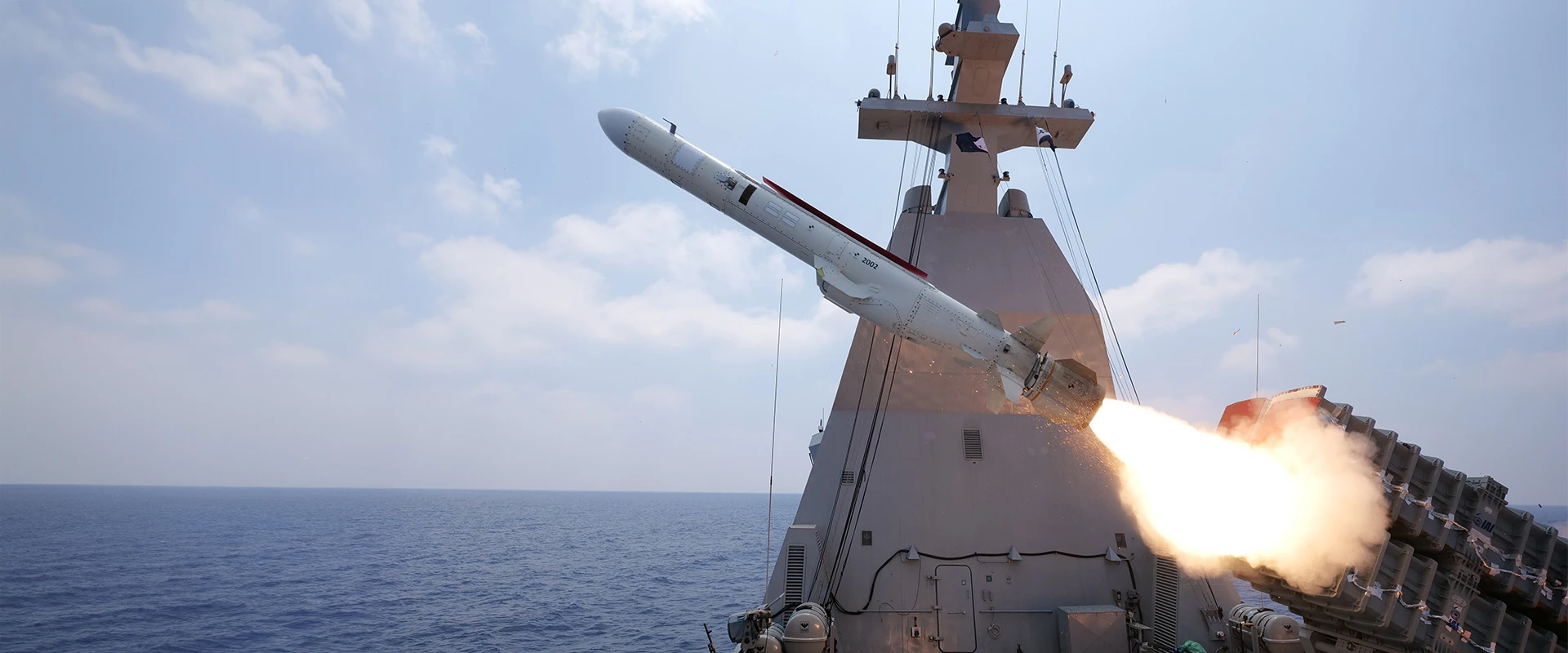A surface to surface missile is being launched from a naval warship over the ocean during daytime, with flames and smoke trailing behind the missile and a clear sky in the background.