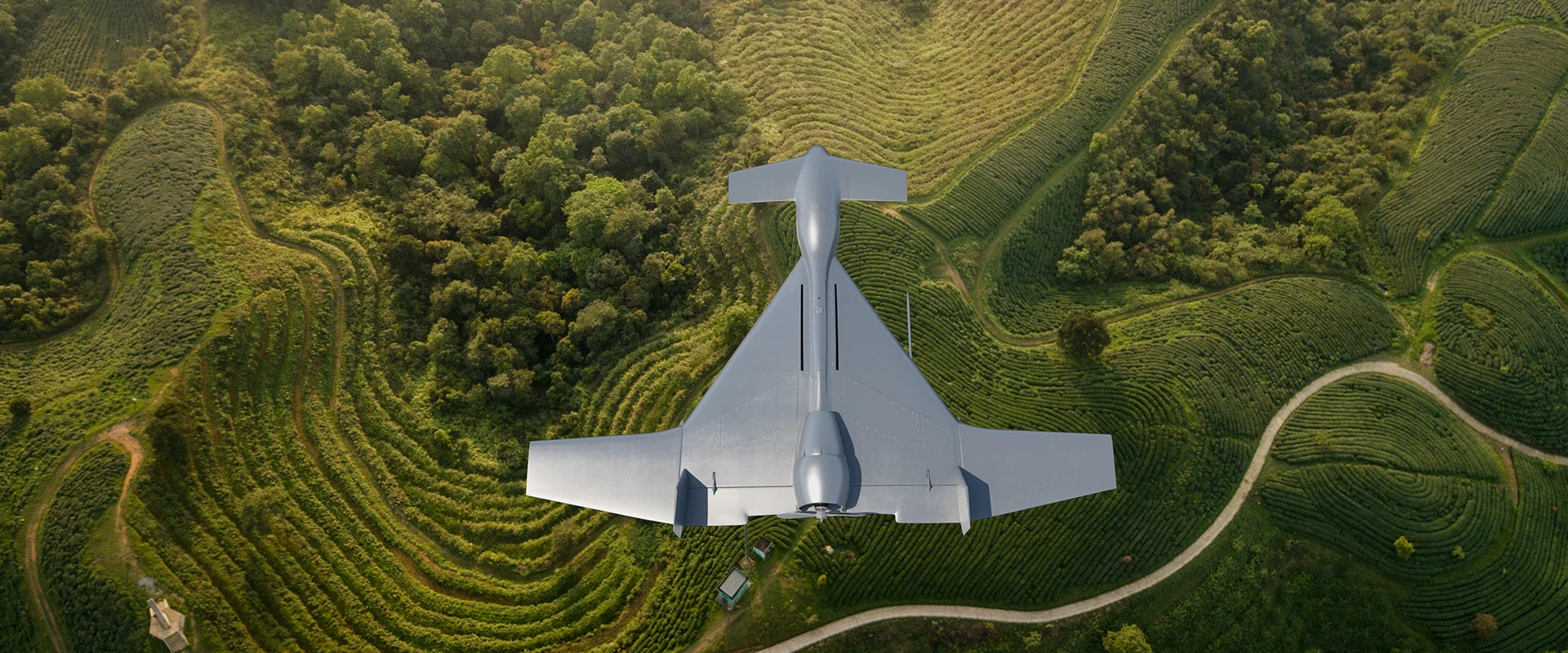 Aerial view of a silver loitering munition system drone flying over green, terraced hills and fields, with a winding road and dense forested area nearby.