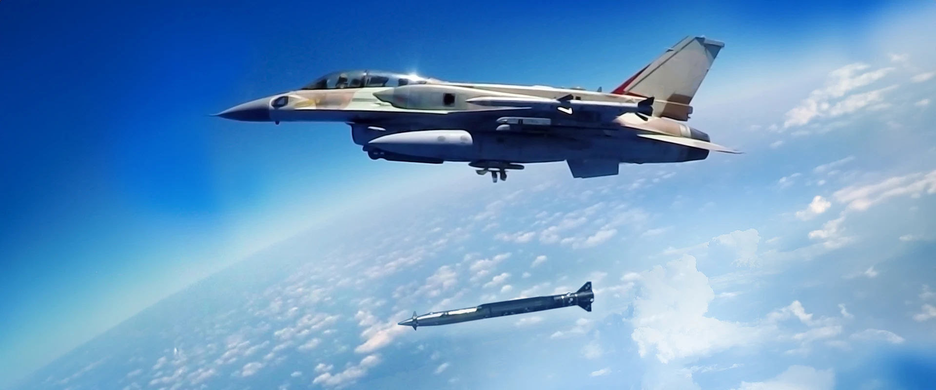 An F-16 fighter jet in flight releases a guided bomb and to ground missile over a partly cloudy sky, with the curvature of the Earth visible in the background.