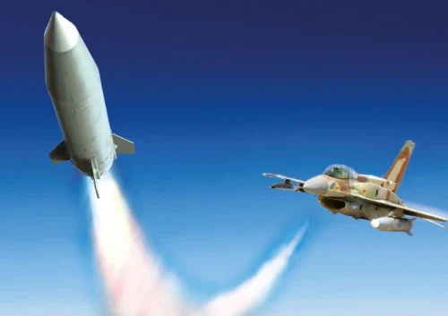 A fighter jet soars through a clear blue sky next to a launched ballistic missile, which ascends upward and leaves a trail of white smoke behind it.