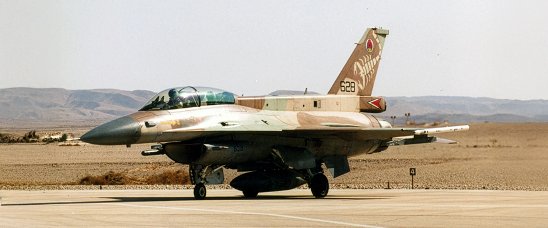 A military jet equipped with a combat training system, sporting desert camouflage, is parked on a runway in an arid landscape. The aircraft has a single vertical tail fin marked with insignia and numbers, with mountains in the background.