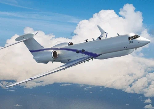A white military jet with a blue stripe and visible antennas, equipped with a signal intelligence system, flies above the clouds against a bright blue sky. The twin-engine jet is marked with the number 002 on its nose.