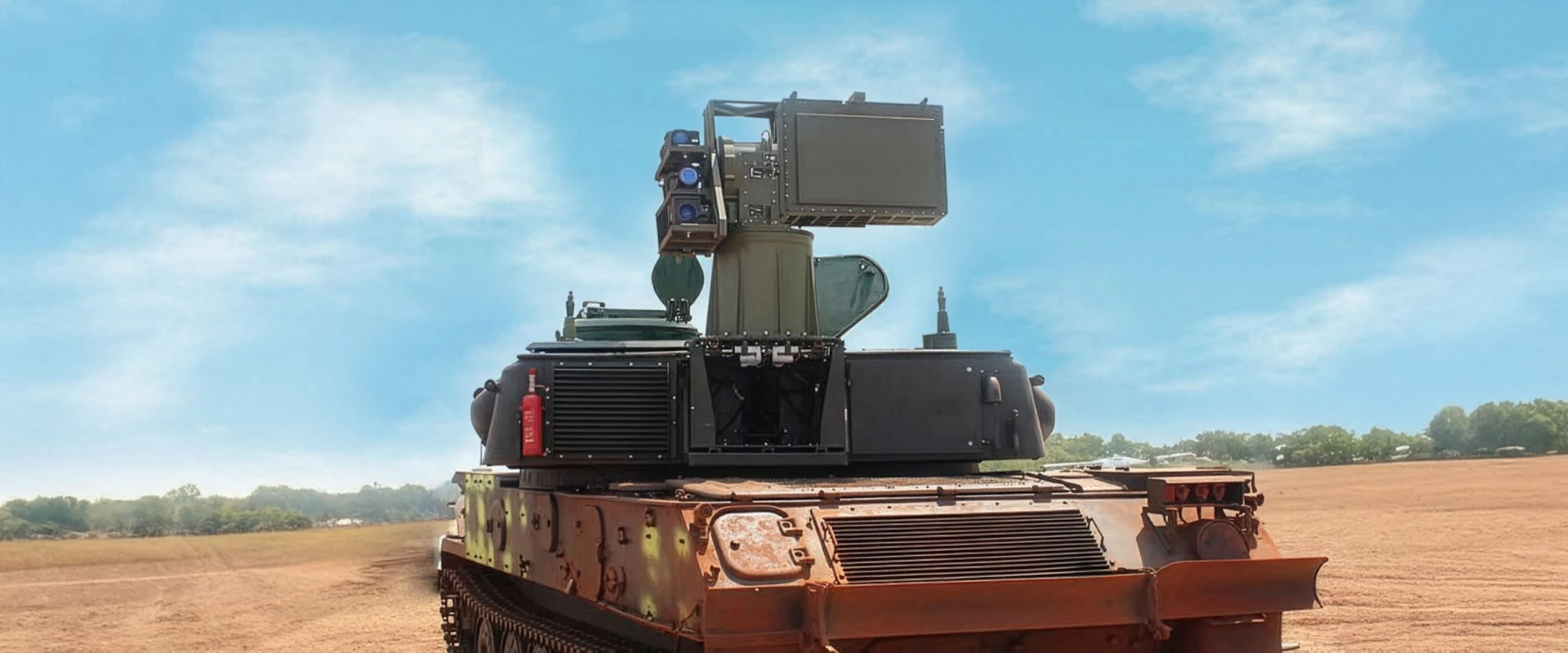 A large military vehicle with mounted radar and track radar equipment is positioned in an open field under a blue sky, with trees visible in the background.