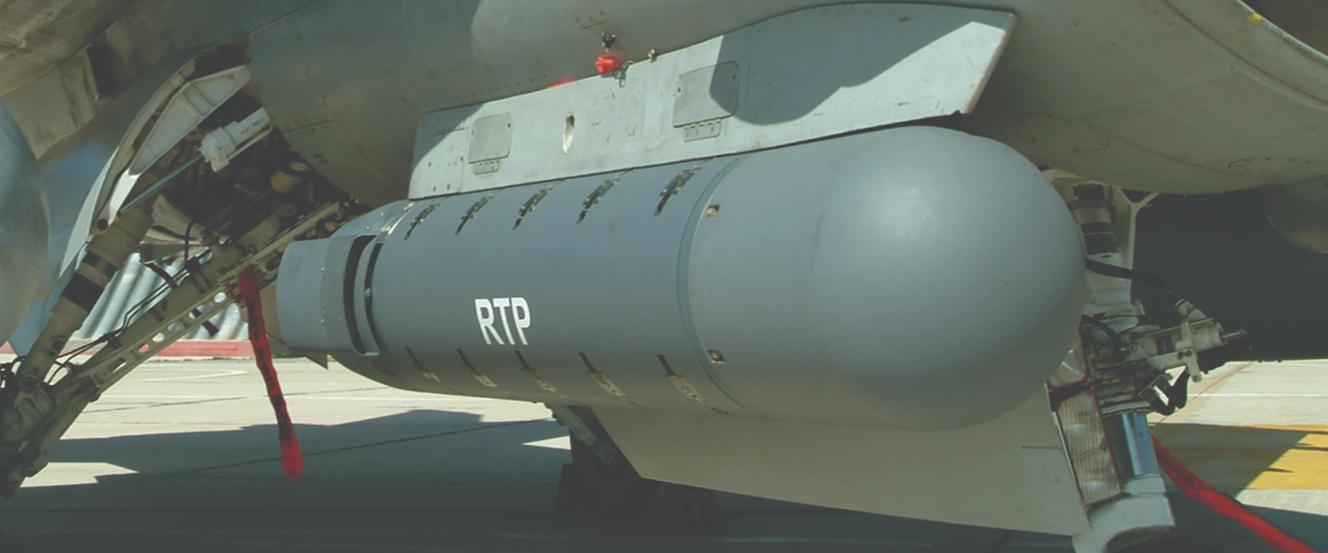 Close-up view of an aircraft’s underbelly showing a large gray external ELM-20600 RTP pod labeled RTP attached near the landing gear, with red safety tags visible nearby.