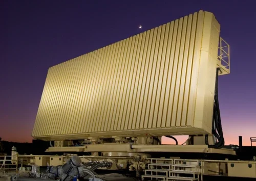 Large ELM 2080C military radar array with vertical panels stands on a platform at dusk, under a purple sky with a crescent moon above. Equipment and railings line the foreground, enhancing the scene’s advanced technological feel.