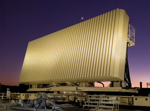 Large ELM 2080C military radar array with vertical panels stands on a platform at dusk, under a purple sky with a crescent moon above. Equipment and railings line the foreground, enhancing the scene’s advanced technological feel.