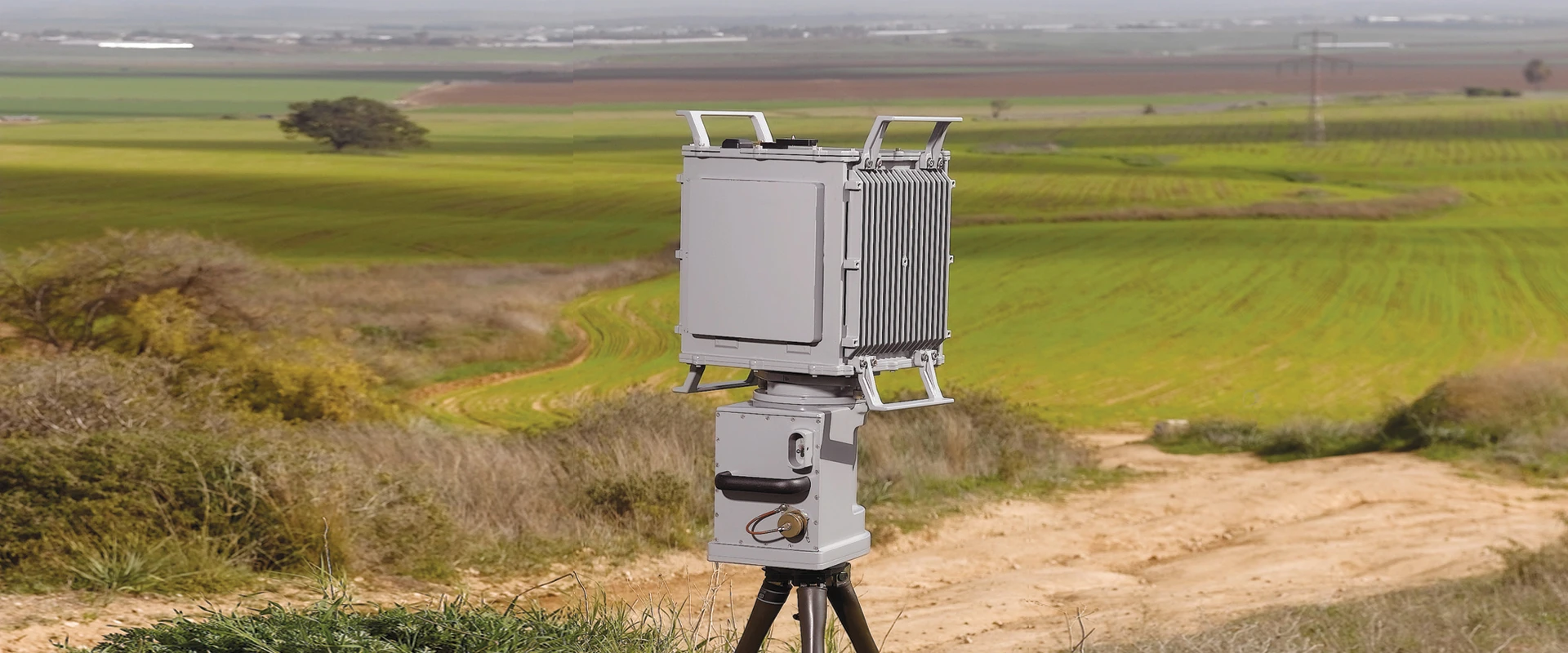 A gray, box-shaped ground surveillance radar is mounted on a tripod in front of a dirt path, surrounded by green grassy fields and patches of farmland under a cloudy sky.