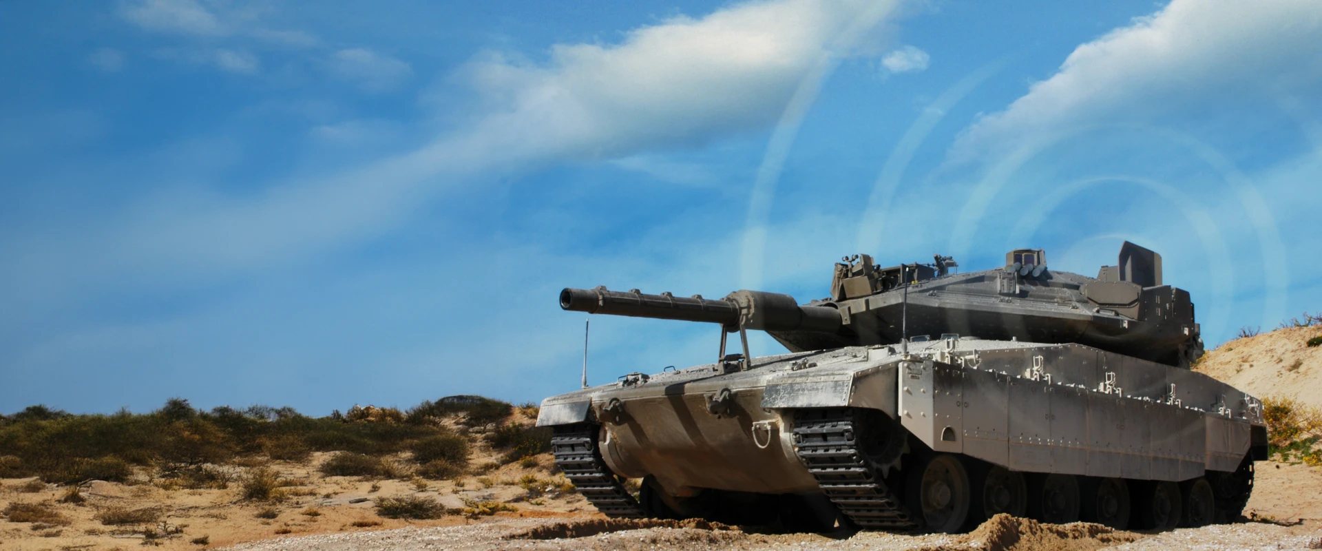 A modern military tank equipped with active protection systems sits on sandy terrain under a blue sky with scattered clouds. Sparse vegetation is visible in the background.