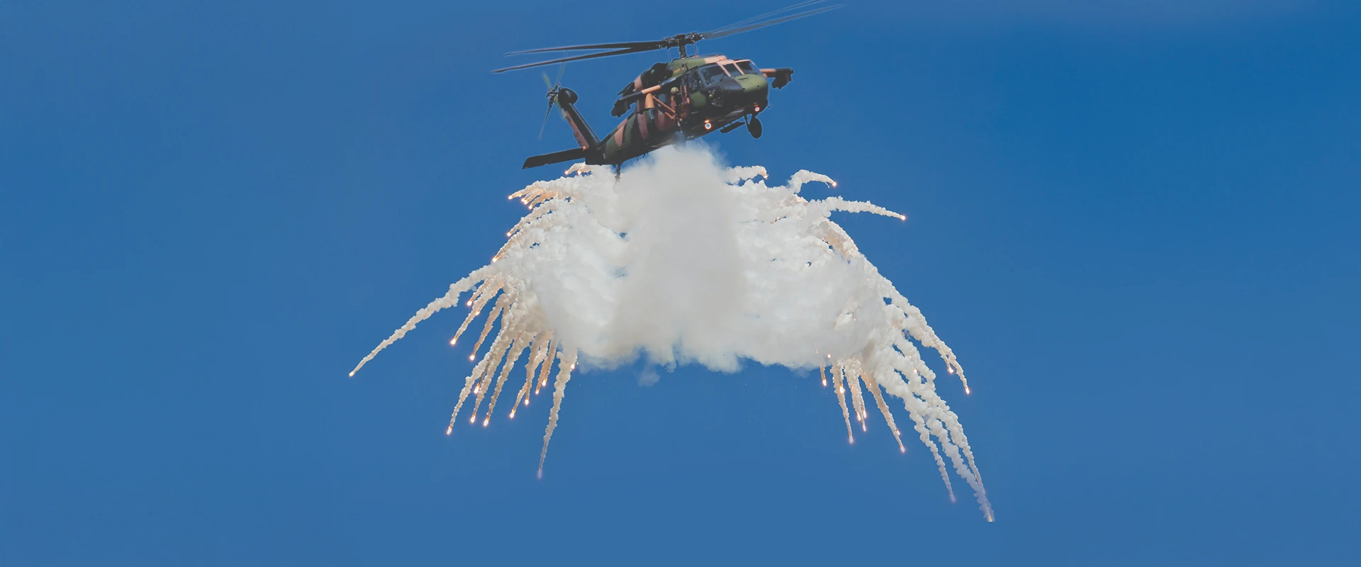 A military helicopter, responding to a missile approach warning, releases flares and smoke in the sky, creating a wing-like effect against a clear blue background.