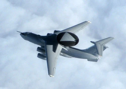 A large military aircraft, the ELW-2090, with four engines and a distinctive radar dome on top, flies above a blanket of clouds.