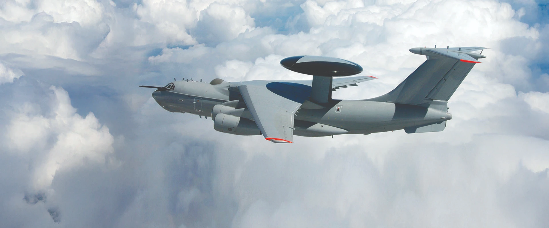 A grey ELW-2090 military aircraft with a large radar dome on top flies above white clouds during the day.