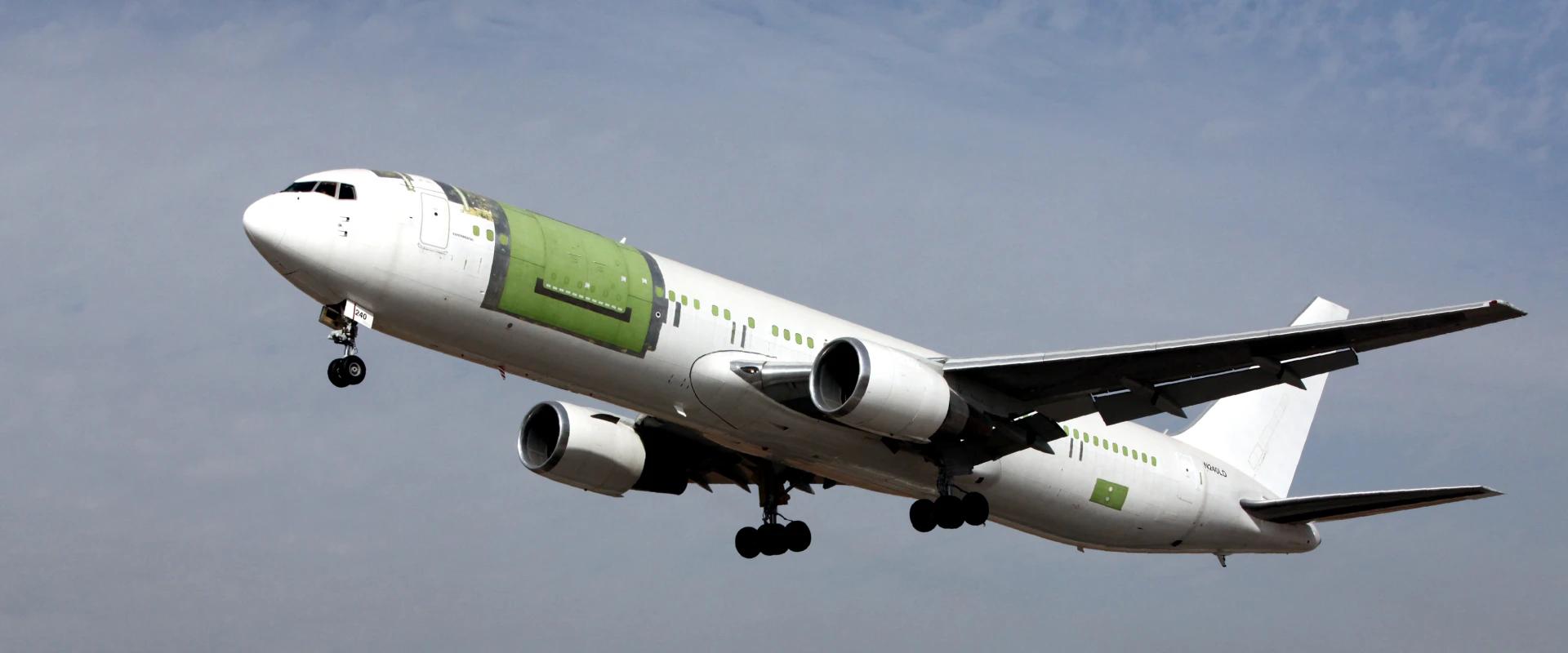 A white commercial airplane with green unpainted panels takes off against a cloudy sky, its landing gear still extended as it ascends during Flight Tests CENTER trials.
