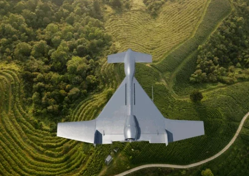 A gray loitering munition system with wide wings flies above green, terraced fields and a winding road, surrounded by dense forest. The aerial view highlights the landscape and the drone’s distinctive shape.