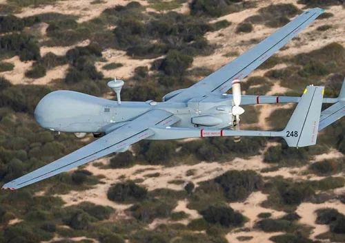 A gray military drone, resembling the Heron MALE UAS with twin tail fins, flies over a desert landscape with sparse vegetation, viewed from above and behind. The number 248 is marked on its tail.