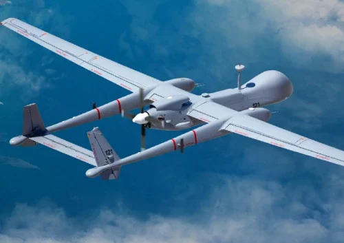 A large gray military drone, resembling the MALE UAV Heron with twin tails and long wings, soars above the clouds, viewed from above against a blue sky backdrop.