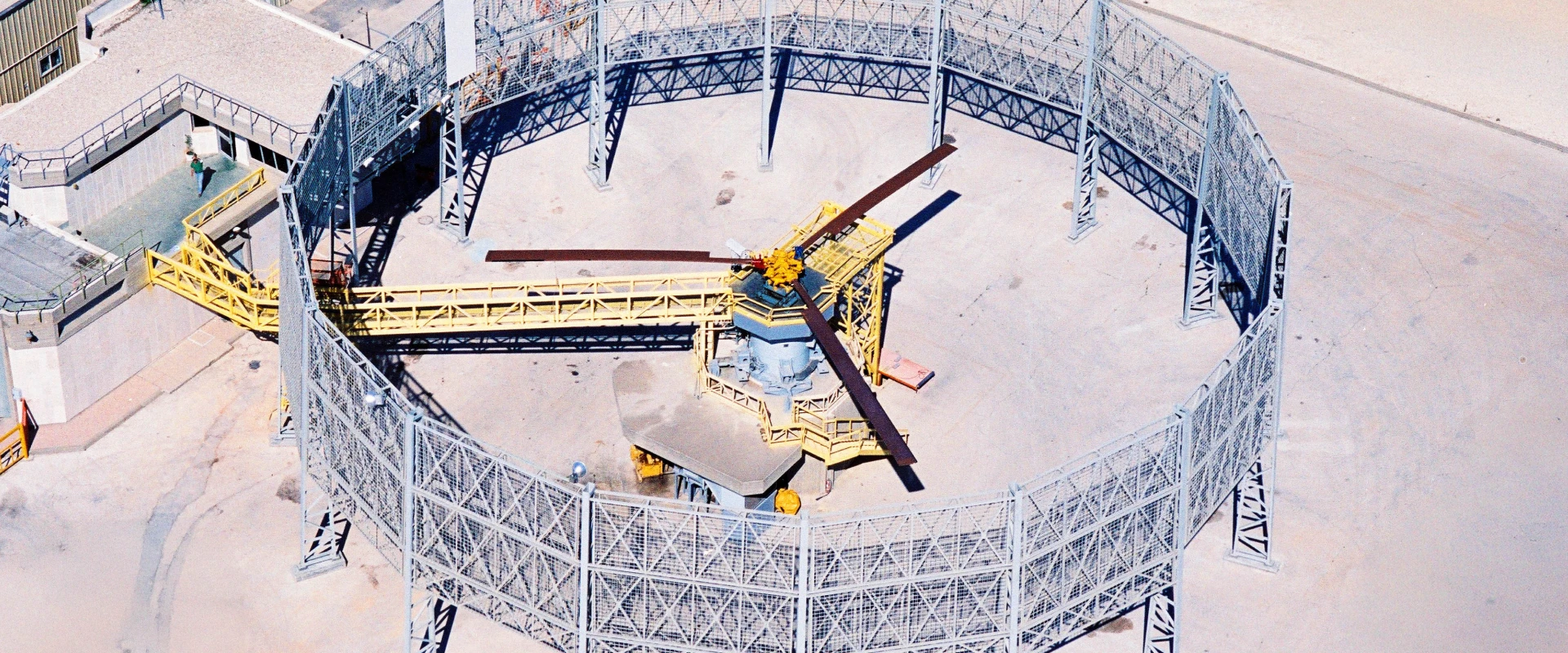 Aerial view of a circular fenced Helicopter MRO facility with a central building and four long beams extending outward, surrounded by sandy terrain. Yellow walkways connect the building to the fence.