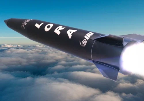 A black precision strike missile labeled LORA flies above the clouds, its engine glowing brightly at the rear against a blue sky.