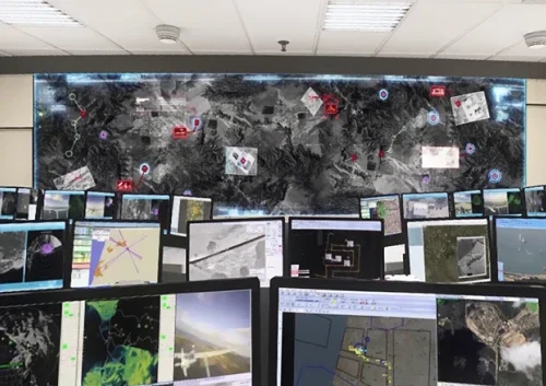 A control room with multiple monitors displaying maps, aerial images, and UAV intelligence data. A large digital map covers the back wall, marked with symbols and routes, indicating a coordinated IAI monitoring or surveillance operation.