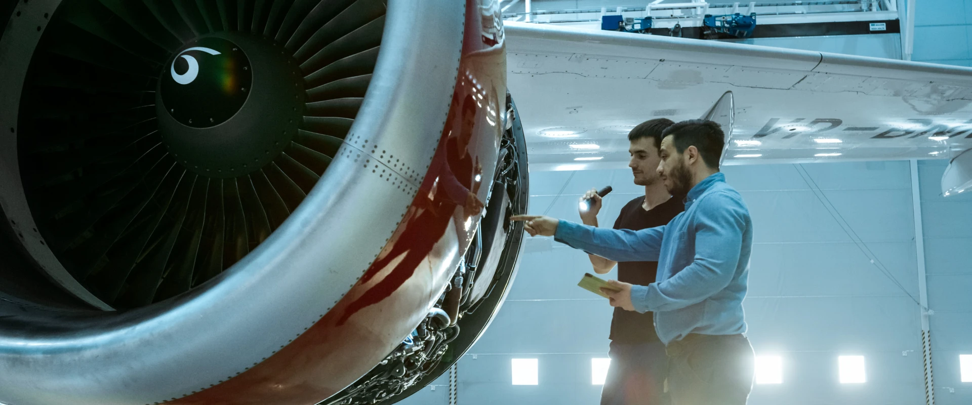 Two men stand near a large jet engine inside a hangar. As they review the engine’s components with RAMS & ILS procedures in mind, one holds a flashlight while the other takes notes, the airplane wing visible above them.