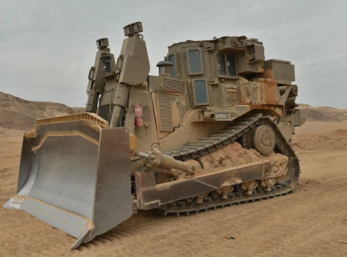A large, armored bulldozer with a robust engineering system and heavy-duty tracks sits on sandy terrain under a cloudy sky, surrounded by barren hills.