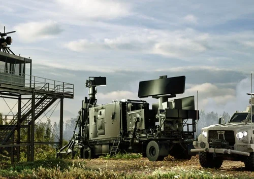 A military hard kill C-UAS radar system mounted on a trailer is positioned outdoors near a metal observation tower and an armored vehicle, under a cloudy sky with trees in the background.