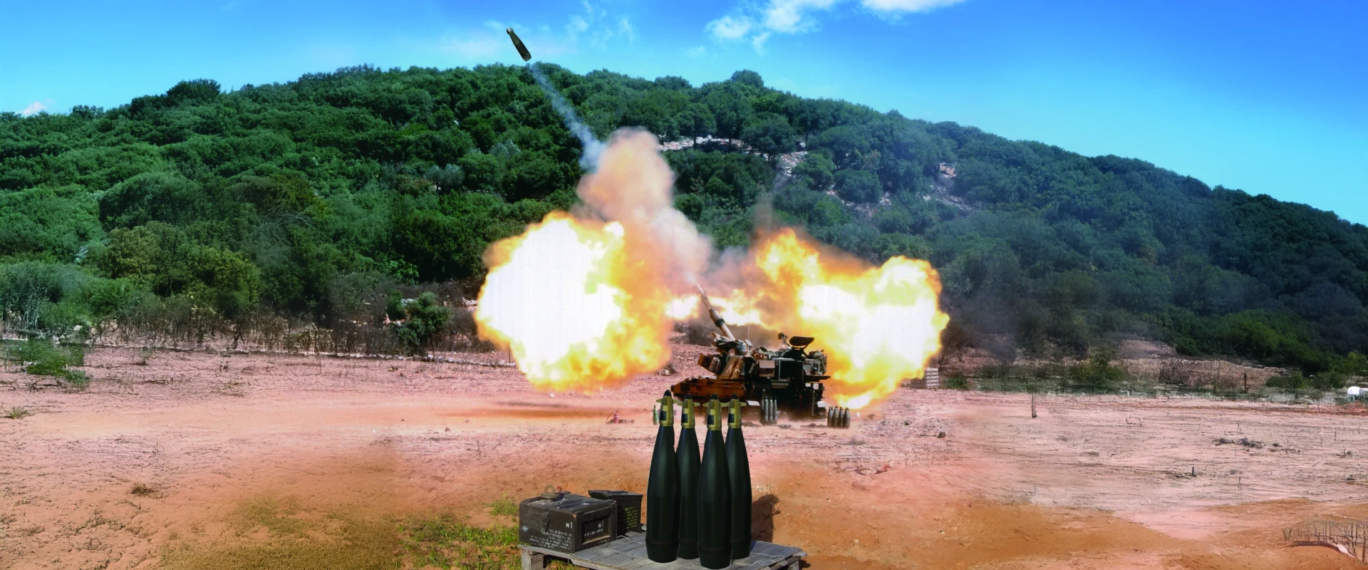 A large artillery gun fires with TOPGUN precision, creating a bright explosion and smoke as a shell soars through the air. Green hills and a blue sky form the backdrop, with several artillery shells visible in the foreground.