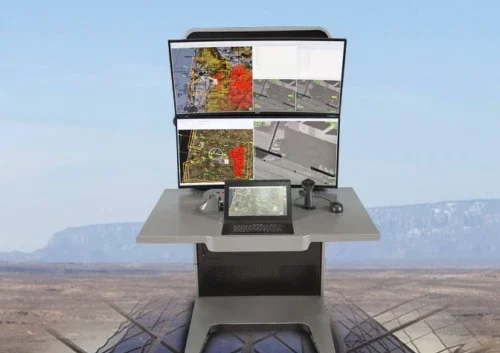A unified control system station with four monitors displaying aerial maps and imagery sits on a desert-like landscape with distant mountains, featuring a keyboard, touchscreen, joystick, and other control devices on the desk.