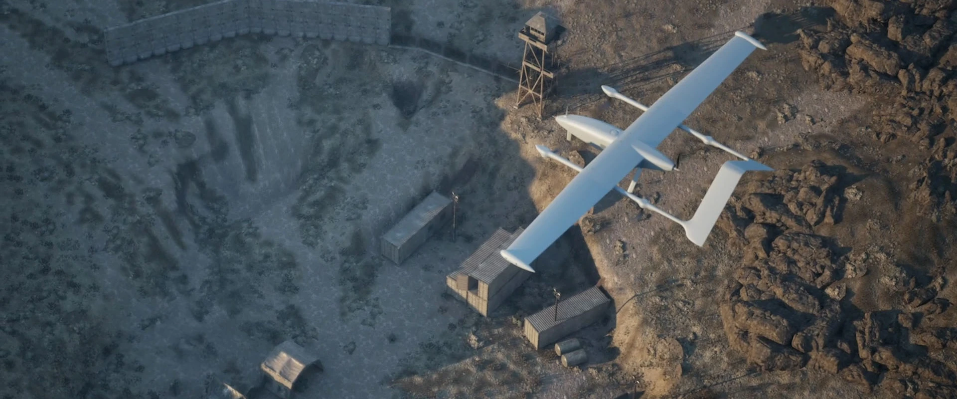 Aerial view of a large white multi-copter UAV flying over a rugged, rocky landscape with scattered shipping containers, small buildings, and a wooden watchtower.