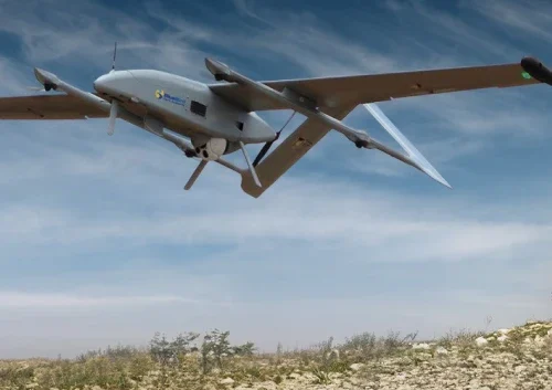 A gray military drone and multi-copter UAV fly low over a rocky landscape with sparse vegetation under a blue sky with wispy clouds.
