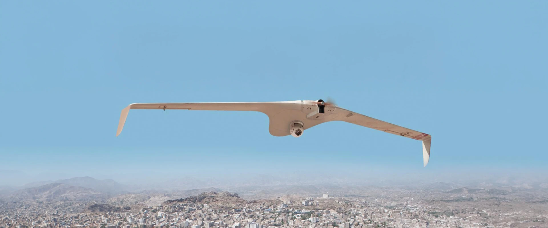 A white, fixed-wing unmanned aerial system flies over a cityscape with mountains in the background and a clear blue sky above.