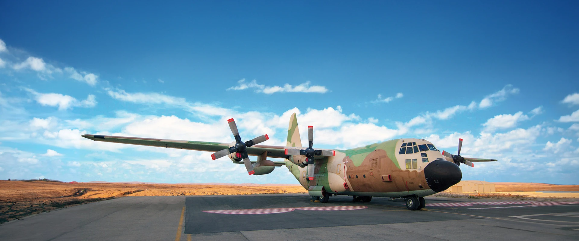 A C130 military transport aircraft with camouflage paint sits parked on a runway under a blue sky with scattered clouds and a desert-like landscape in the background.