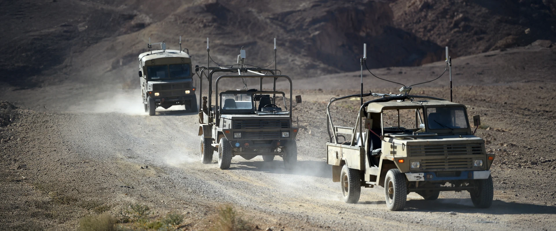 Three military vehicles drive in a single file along a dusty, rocky road in a desert landscape, likely as part of a logistics support system. Dust clouds rise behind them, and barren hills stretch beneath the clear sky.