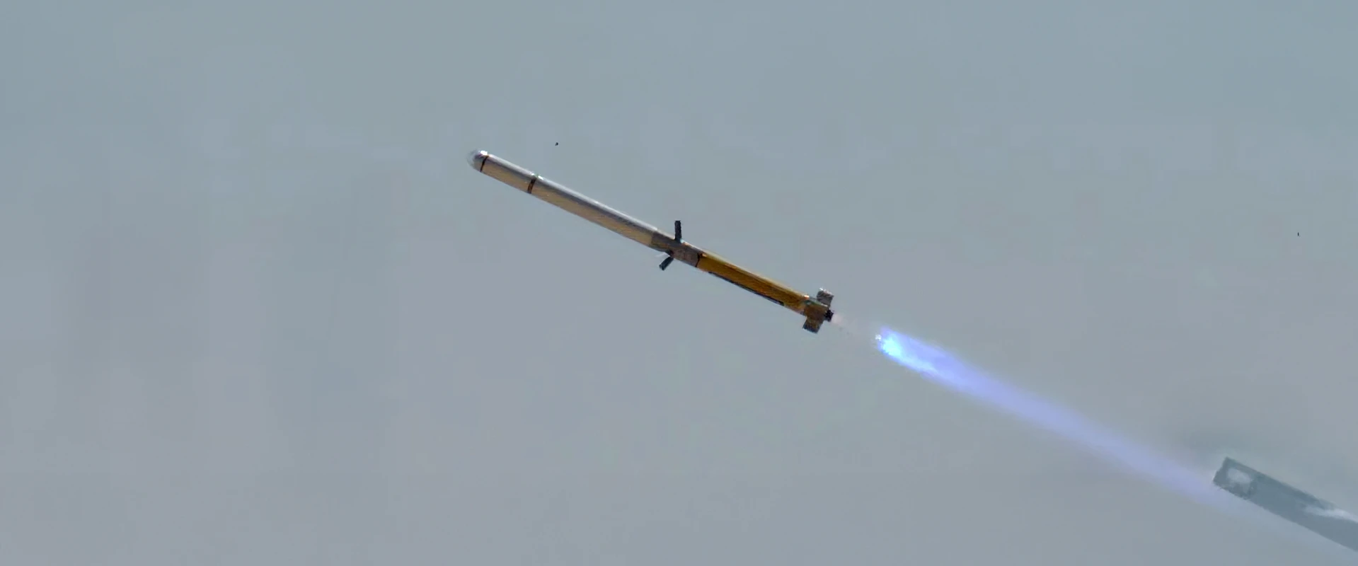 A rocket, resembling an anti tank missile, flies through the sky at an angle, producing a bright blue flame from its engine against a pale, cloudless background.
