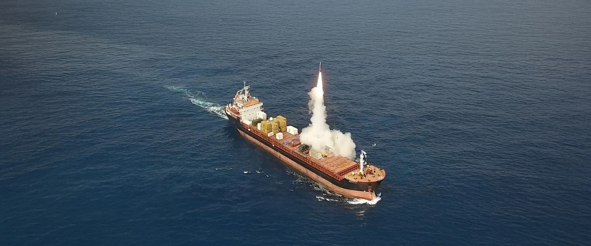 A cargo ship at sea launches a Naval LORA missile vertically from its deck, smoke trailing behind as it ascends into the sky. The ship is surrounded by calm blue water.