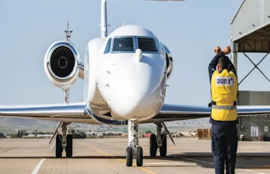 A ground crew member in a yellow vest signals to a white private jet as it taxis on the runway, demonstrating key ground crew focus near a hangar at an airport.
