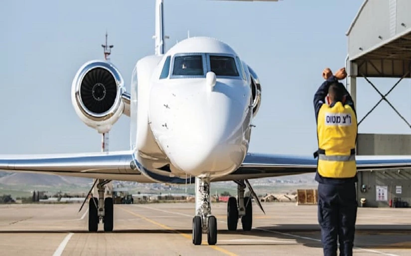 A ground crew member in a yellow vest signals to a white private jet as it taxis on the runway, demonstrating key ground crew focus near a hangar at an airport.